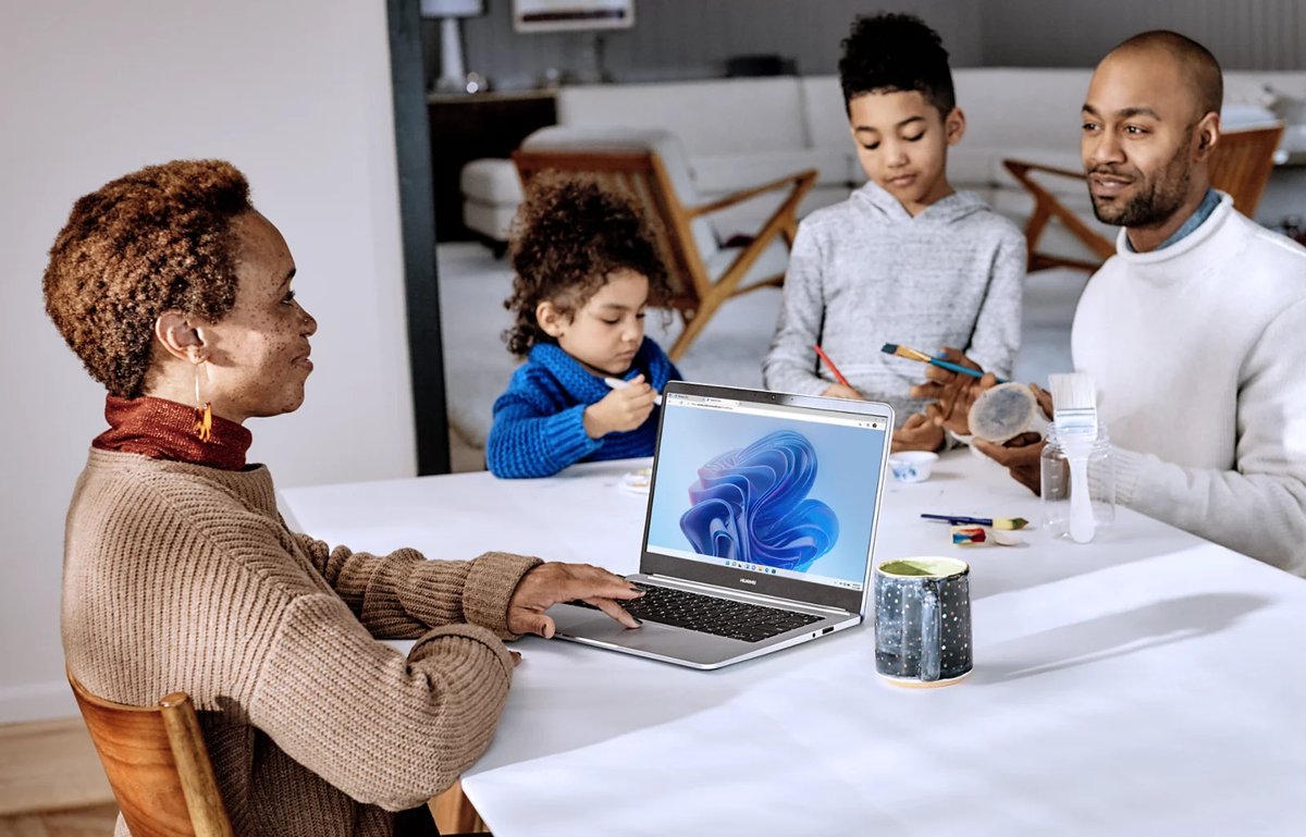 An adult using a Windows 11 laptop at a dining table while two children paint alongside